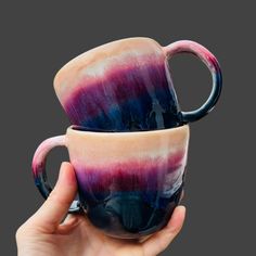 two handmade coffee mugs with purple and blue swirled designs on the sides, held in front of a gray background