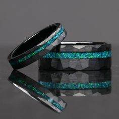 two black rings with green and blue glitter inlays are shown on a reflective surface