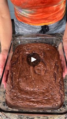 a person holding a cake in a pan on top of a granite counter with an orange pumpkin behind it