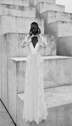 a woman in a long white dress is standing on concrete steps and talking on the phone