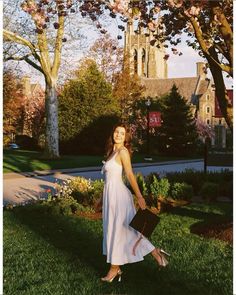 a woman in a white dress holding a briefcase and standing on the grass near some trees