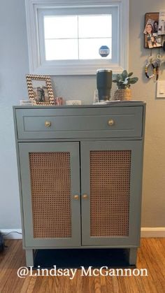 a gray cabinet with wicker doors and drawers in front of a window, next to a wooden floor