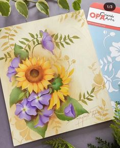 a card with flowers painted on it next to some leaves and plants in the background