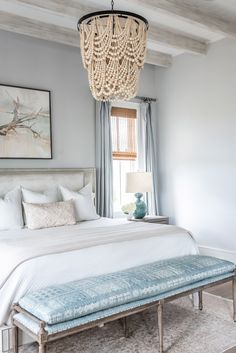 a white bed sitting under a chandelier next to a window in a bedroom