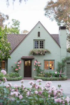 a green house with pink flowers in the front yard and windows on each side of the house