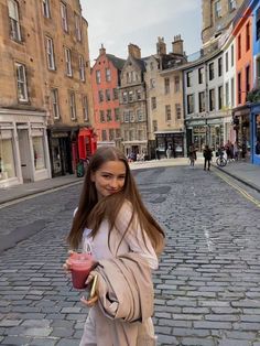 a girl walking down the street holding a drink in her hand and looking at the camera