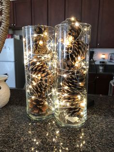 pine cones are placed in clear glass vases on the counter with fairy lights around them