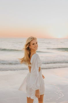 a beautiful blonde woman standing on top of a beach next to the ocean at sunset