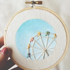 a person holding up a small embroidery art piece with a ferris wheel in the middle