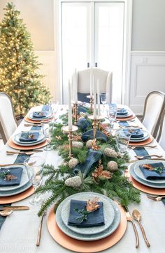 a dining room table set for christmas with blue and gold place settings, silverware, candlesticks and greenery