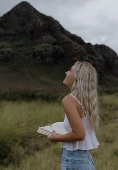 a woman standing in a field holding a book and looking up into the sky with a mountain in the background