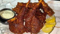 a plate full of ribs with dipping sauce and lemon wedges next to it on a marble table