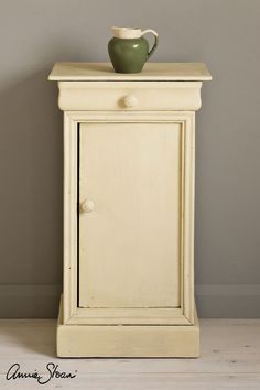 a small cabinet with a green vase on top and a white wall in the background