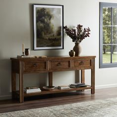 a wooden table with two drawers in front of a painting on the wall next to a window