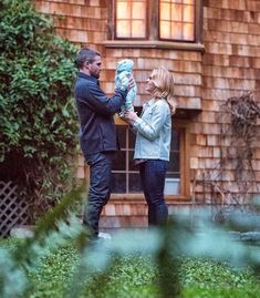 a man standing next to a woman in front of a brown house holding a baby