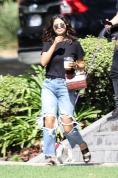 a woman in ripped jeans and black shirt walking her dog down the street with a drink