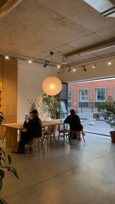 two people sitting at a table in an open room with large windows and potted plants