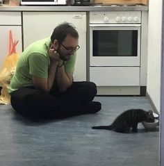 a man sitting on the floor next to a black cat eating out of a bowl