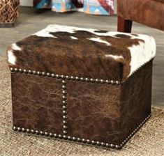 a brown and white cowhide ottoman with studded trim on the top, sitting on a rug in front of a chair