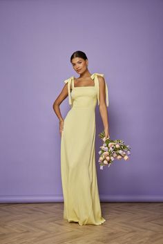 a woman standing in front of a purple wall wearing a yellow dress and holding a bouquet