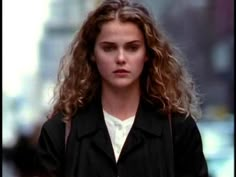 a woman with long curly hair walking down the street wearing a black jacket and white shirt