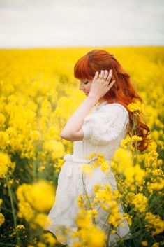 a woman with red hair standing in a field of yellow flowers holding her hand to her head