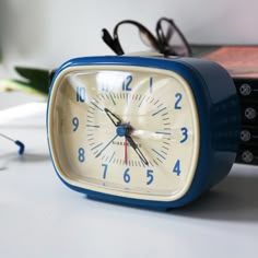 a blue alarm clock sitting on top of a table next to eyeglasses and a book