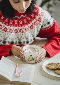 a woman sitting at a table in front of an open book holding a mug with marshmallows on it