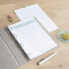 an open binder next to two notebooks on top of a wooden table with pens and paper clips