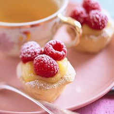 two small pastries with raspberries on them sit on a pink plate next to a cup of tea