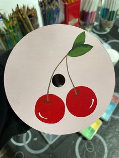 a person holding up a paper plate with two cherries on it and paintbrushes in the background