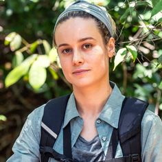 a woman wearing a headband standing in front of some trees and bushes, looking at the camera