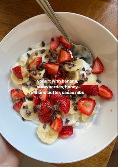 a bowl filled with bananas and strawberries on top of a wooden table next to a hand holding a spoon