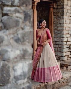 a woman in a pink and beige lehenga standing by a stone wall with her hands on her hips