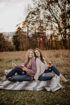 two women sitting on a blanket in the middle of an open field, drinking beverages