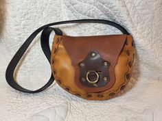 "This leather bag is made from a 1970's pattern that we dug up, it has a cool retro feel but also works well for steampunk or modern style! It is made of sienna brown bull hide, a goldish tan cowhide, and dark brown cowhide front detail with black latigo for the strap.  It is completely hand stitched with our own very strong leather cording that we make, so this is a very sturdy bag.  This one has a decorative piece on the front flap with antiqued rivets and a slot which has a brass ring that passes through to hold it closed.  The bag is 9\" wide at the widest point, 8\" tall, and the bottom is about 3.5\" deep. The strap is 2\" wide and 38\" long, can be worn on the shoulder or across the body." Cognac Satchel Shoulder Bag With Brass Hardware, Brown Satchel Shoulder Bag With Brass Hardware, Brown Crossbody Shoulder Bag With Brass Hardware, Brown Shoulder Satchel With Brass Hardware, Brown Satchel With Brass Hardware, Brown Saddle Shoulder Bag With Brass Hardware, Brown Satchel Bags With Brass Hardware, Brown Satchel With Brass Hardware Shaped As Tote, Gold Leather Saddle Bag For Everyday Use