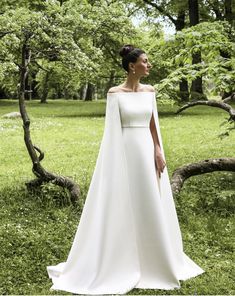 a woman in a white wedding dress standing in the grass with her cape over her shoulders