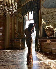 a woman in a long dress is standing by a chandelier and looking into the mirror