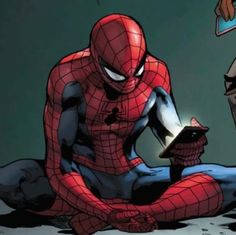 a spider - man sitting on the ground next to another person holding an electronic device