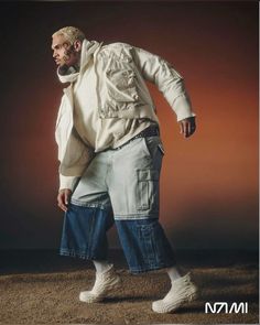 a man in white jacket and jeans standing on dirt ground with his hands behind his back