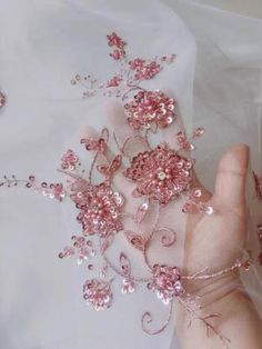 a hand is holding some pink flowers on a white cloth with beads and sequins