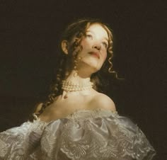 a woman with curly hair wearing a white dress and pearls on her neck, looking up