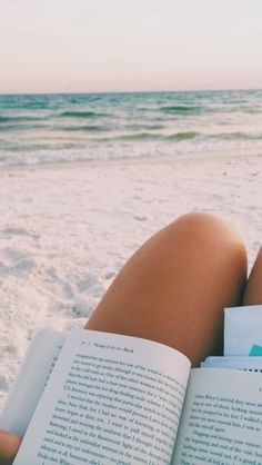 a woman reading a book on the beach with her feet in the sand and water behind her