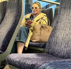 a woman sitting on a train looking at her cell phone while holding a purse and wearing sunglasses