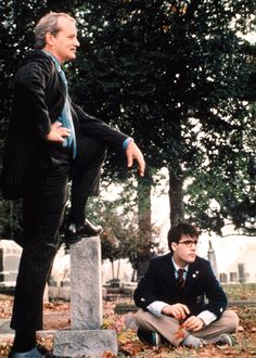 two men in suits sitting on tombstones with one pointing at the other man standing