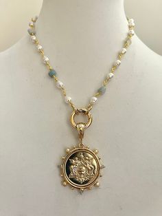 An elegant pearl and aqua stone rosary chain necklace with gold wire and a French Le Monde replica coin pendant. The pendant hangs off a beautiful thick gold bale. The coin bezel, an Art Deco French stunner has Clear CZ and Pearl accents. The clasp is a spring ring and so easy to handle. Please see the picture of the clasp opening. Material: Bezel is made of Cast Zinc Alloy and the coin is of brass Measure - Bezel and coin = 40mm Coin = 28mm Spring ring =20mm Tips to keep your jewelry looking go Gold Pearl Jewelry With Coin Pendant, Gold Medallion Jewelry With Pearl Charm, Gold Medallion Pearl Necklace With Pendant, Gold Pearl Pendant Necklace In Medallion Shape, Gold Pearl Necklace With Medallion Pendant, Gold Pearl Necklace With Coin Pendant, Gold Medallion Jewelry With Pearl Chain, Gold Coin Necklace With Pearl Chain, Rosary Chain Necklace