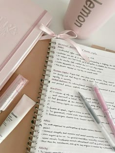 a pink notebook and pen sitting on top of an open notepad next to a cup