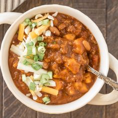 a white bowl filled with chili and cheese on top of a wooden table next to a spoon