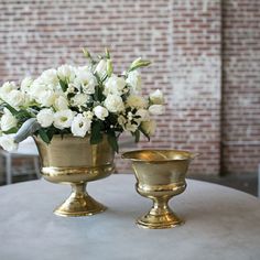 two gold vases with white flowers in them on a table next to a brick wall