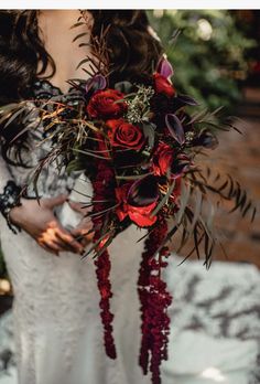 a woman in a wedding dress holding a red bouquet with greenery and flowers on it